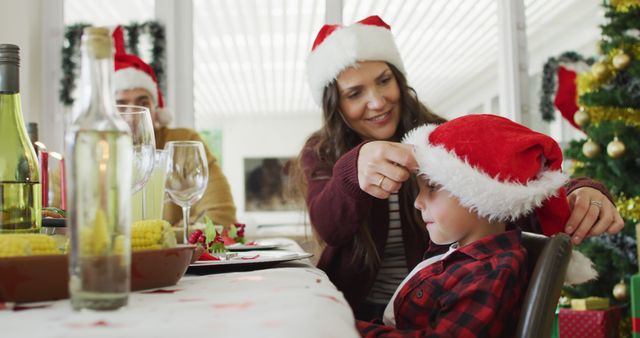 Mother helps her young child adjust his Santa hat at a holiday dinner table adorned with festive decorations. Perfect for representing family warmth, festive celebrations, holiday greetings, and togetherness during the Christmas season. Can be used in advertisements, holiday cards, family blogs, social media posts, and winter holiday promotions.