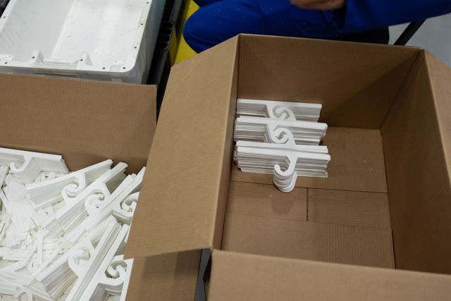 High angle front view of a pile of white, plastic hangers being stacked neatly in an open cardboard box in a factory warehouse, in preparation for shipment