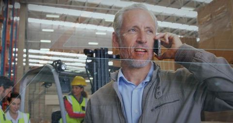 Warehouse manager talking on phone behind glass partition with forklift and team in busy warehouse