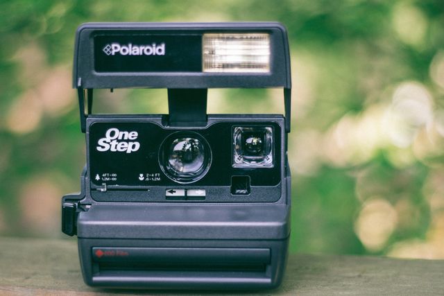 Vintage Polaroid OneStep camera positioned outdoors, with a blurred green nature background. Ideal for use in content about retro photography, nostalgic equipment, or vintage tech reviews. Great for illustrating articles or blog posts about the history of cameras, the resurgence of analog photography, or the aesthetic appeal of old-fashioned items.