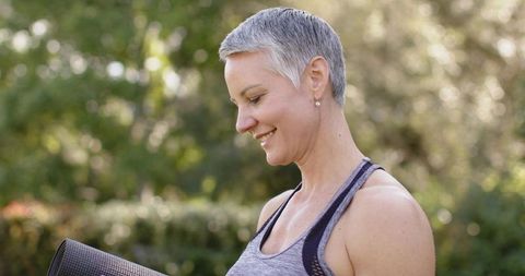 Mature woman holding yoga mat smiling during outdoor yoga session in sunlit garden