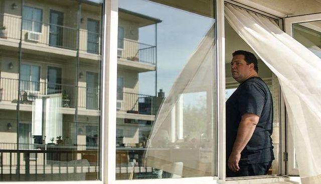 Man standing at sliding glass door viewing urban balcony reflection, interior daylight