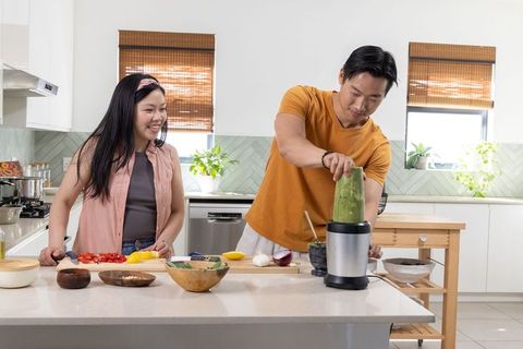 Asian couple preparing green smoothie and chopping veggies in bright modern kitchen