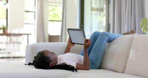 Teen african girl lounging on beige sofa using tablet in bright modern open-plan living room