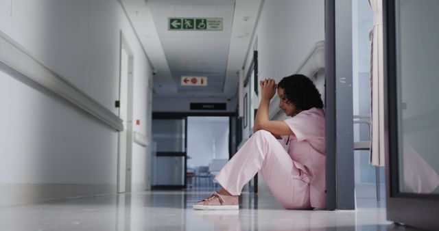 A nurse in pink scrubs is sitting on the floor of a hospital corridor, looking exhausted and stressed. Use this to represent the challenges and mental health struggles faced by healthcare professionals, especially during demanding times. Ideal for articles, blogs, or campaigns related to medical professions, mental health awareness, and nurse appreciation.