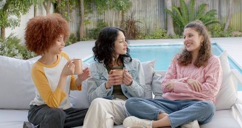 Diverse women relaxing on poolside sofa, chatting, drinking coffee, enjoying outdoor lounge