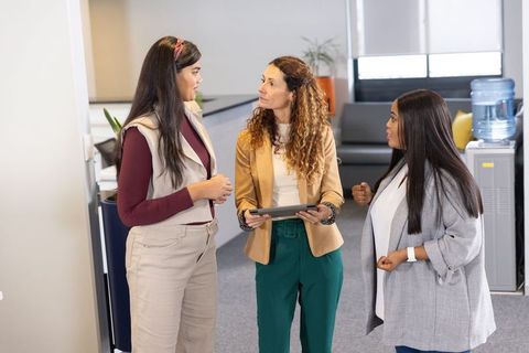 Diverse female coworkers discussing project while holding tablet in open-plan office