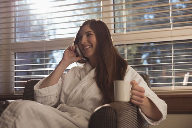 Woman sitting in living room, wearing a robe, talking on mobile phone while holding a coffee mug. Ideal for themes related to home comfort, morning routines, leisure activities, and communication. Suitable for lifestyle blogs, advertisements for home products, or articles about relaxation and self-care.