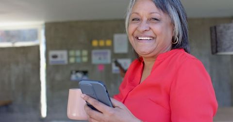 Smiling senior enjoying device while holding mug in office