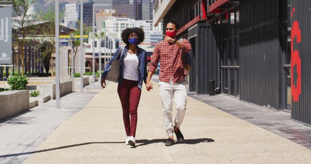Couple strolling in an urban area while wearing protective masks, holding hands and enjoying time together. Suitable for content related to COVID-19 safety, pandemic lifestyles, urban love stories, or promoting social distancing measures.