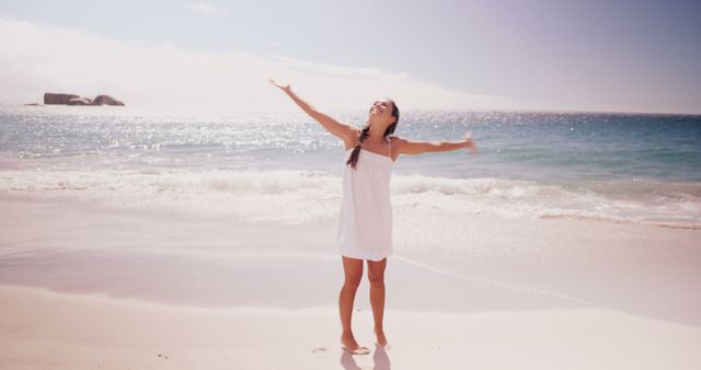 A young Caucasian woman enjoys the sunshine with her arms outstretched on a sandy beach, with copy space. Her expression of freedom and joy encapsulates the essence of a carefree summer day by the sea.