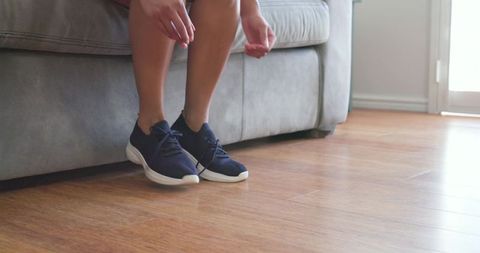Woman sitting on sofa reaching for navy sneakers on hardwood floor near sliding door