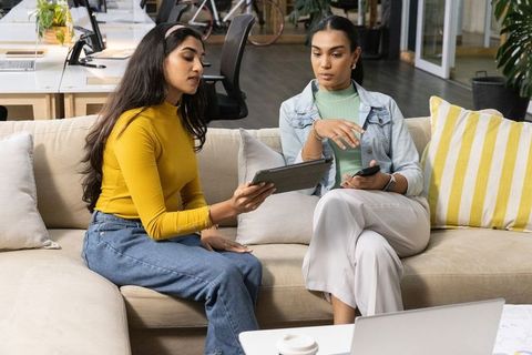 Diverse female coworkers collaborating on tablet and phone in modern office lounge