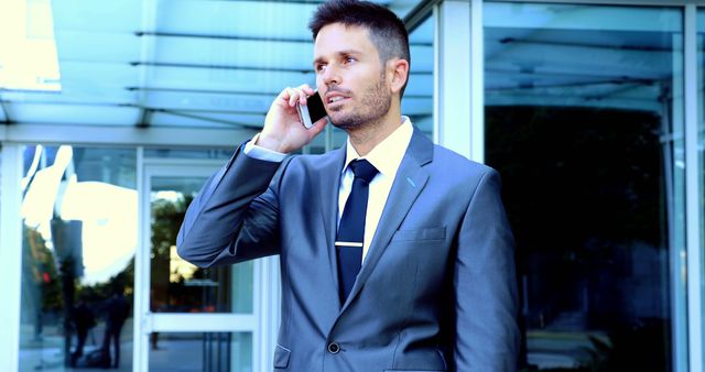 Confident businessman in suit and tie talking on smartphone outside modern office building. Suitable for topics related to corporate communication, business professionalism, executive roles, and business conduct.