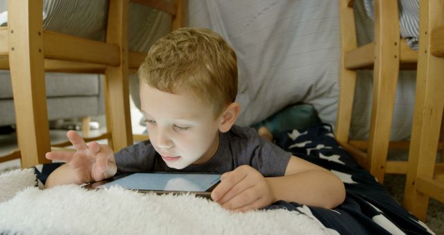 Young boy engaging with a tablet while lying in a cozy blanket fort at home. Bright and cozy atmosphere creates a feeling of warmth and comfort. Perfect for illustrating concepts related to childhood, technology, indoor activities, and family bond. Suitable for blogs, articles, educational content, and advertisements focusing on children, digital learning, and indoor playtime.