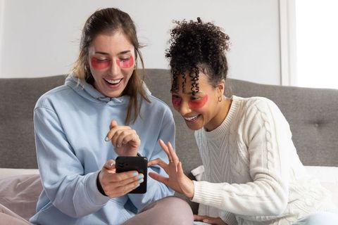Friends sharing skincare moment on bed laughing and browsing phone