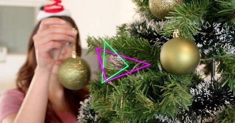 Woman hanging gold ornament on christmas tree at home with santa headband and garland
