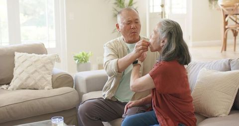 Asian senior man helping partner take medication on sunlit sofa, caregiving and calm