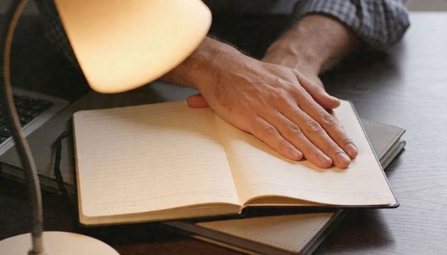 Man Resting Hand on Open Lined Notebook Wearing Checked Shirt at Desk Lamp Laptop Planner
