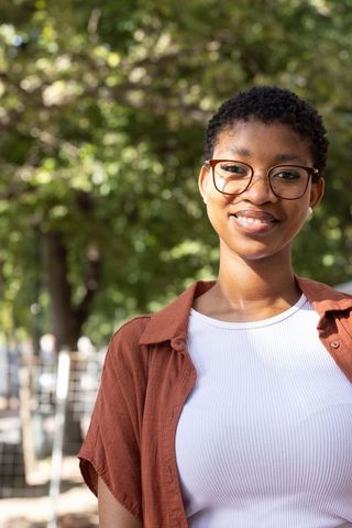 Sunlit african american woman standing and smiling on park path wearing glasses white top rust shirt