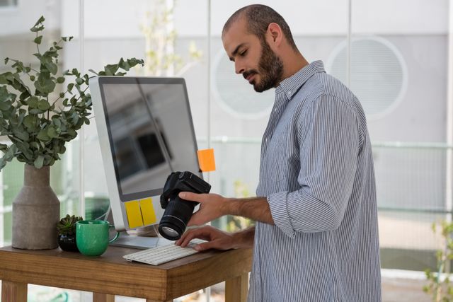 Designer examining camera while standing by computer in a modern studio. Ideal for use in articles or advertisements related to photography, creative workspaces, professional designers, or modern office environments. Can also be used in blogs or websites focused on technology, digital equipment, or creative professions.