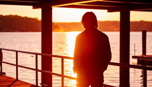 Silhouette man standing on covered pier at golden sunset over lake, quiet reflection