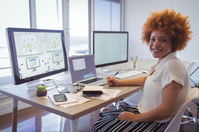 Businesswoman sitting at desk in modern office, working on multiple screens. Ideal for use in business, technology, and productivity contexts. Suitable for illustrating modern work environments, professional women, and creative industries.