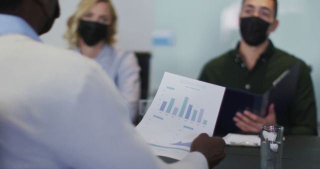 Three business professionals, wearing masks, are discussing a financial report with bar graphs during a meeting. Use this image to illustrate corporate presentations, team collaborations, financial analysis, data presentation, and business strategy meetings.