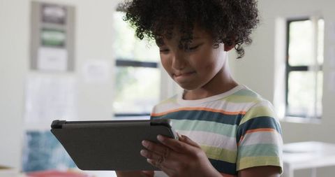 Curly-haired boy learning on tablet and focusing in bright classroom, student technology