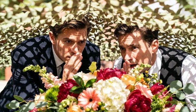 Two men in suits whispering over wedding bouquet under camouflage netting with patterned shadows