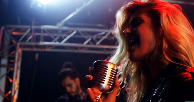 A Caucasian female singer passionately performs into a vintage microphone on stage, with a male band member in the background, with copy space. Her dynamic expression and the stage lighting capture the energy of a live music performance.
