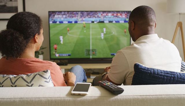 Couple watching soccer match on tv from sofa with remote and smartphone, cozy living room