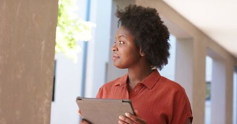 African woman standing in modern corridor holding tablet and gazing at sunlit greenery