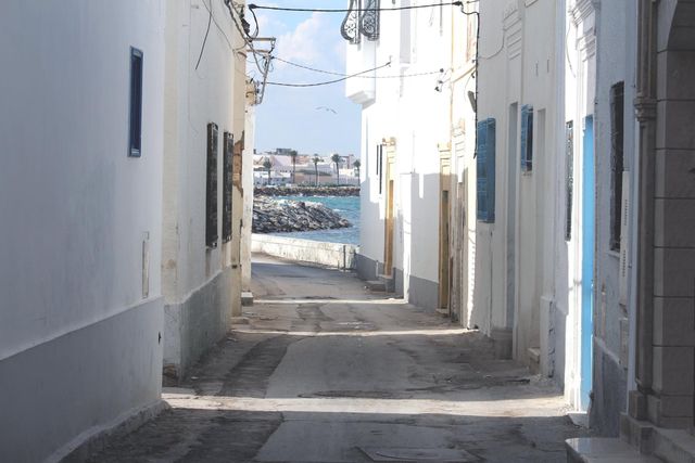 Narrow street with white buildings on both sides, leading to the Mediterranean Sea. Sunlight creates a bright and inviting atmosphere. Ideal for use in travel brochures, articles about Mediterranean towns, or images promoting tourism to seaside destinations.