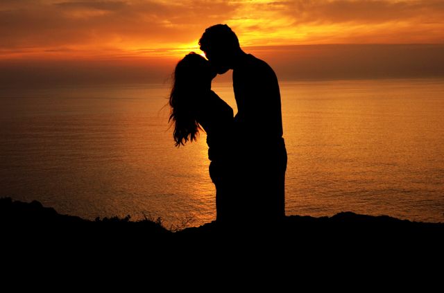 Silhouette of a couple kissing against a breathtaking sunset sky over the ocean. The warm glow of the sunset casts a romantic ambiance, perfect for depicting themes of love and romance. Ideal for use in relationship blogs, romantic greeting cards, travel brochures, and dating website imagery.