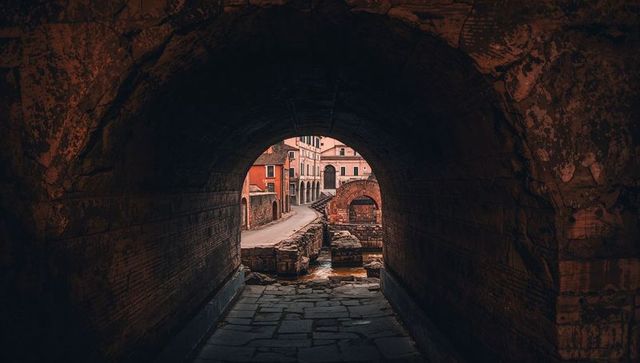 Vaulted stone tunnel leading to medieval riverside street with arched bridge