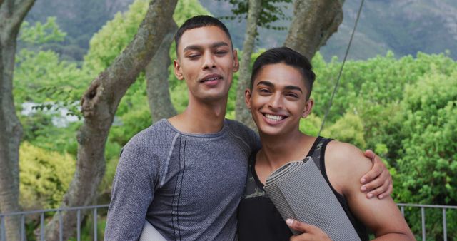 Two friends standing outdoors, each holding a yoga mat, smiling, and enjoying time together. The scene includes green trees and mountains in the background, suggesting this may be in a park or nature area. Perfect for use in content related to outdoor activities, healthy lifestyles, fitness, and building friendships.