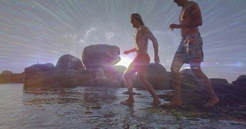 Two men walking on rocky tidepool at sunset with sunburst flare and coastal reflections