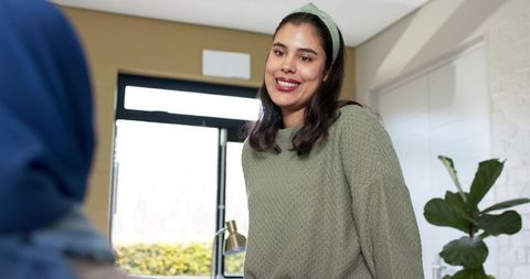 Indian woman smiling and chatting with friend in cozy sunlit living room near glass door