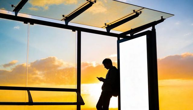 Silhouette commuter checking smartphone at glass bus shelter during golden hour sunset