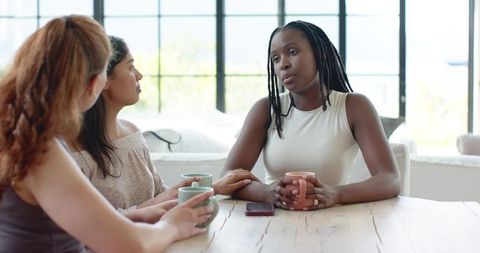 Three women having heartfelt conversation while holding coffee mugs at kitchen table