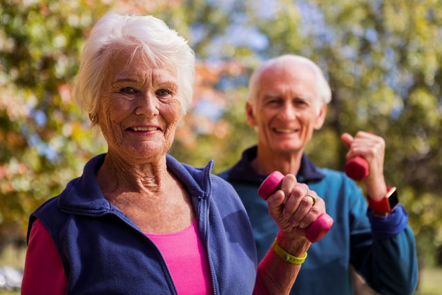 Elderly couple lifting dumbbells and smiling in a park during autumn. Ideal for promoting senior fitness, healthy aging, outdoor activities, and wellness programs. Suitable for use in health and fitness articles, retirement community brochures, and advertisements for senior exercise classes.