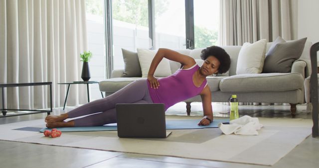 Woman performing a side plank from an online workout in her living room. She is using a laptop to follow along. Water bottle, red dumbbells, and a towel are nearby. Great for promoting home fitness, healthy living, online workout programs, and lifestyle blogs about exercise routines.