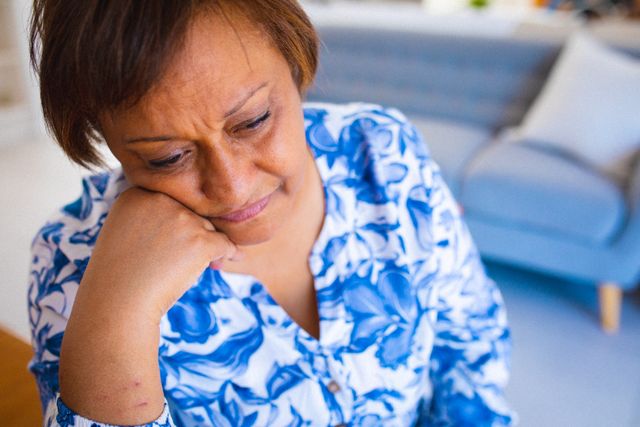 This image depicts a sad African American senior woman sitting at home with her head in her hand. It can be used in articles or campaigns related to mental health, elderly care, loneliness, depression, retirement, and emotional well-being. It is suitable for illustrating the challenges faced by seniors, especially in the context of isolation and mental health issues.