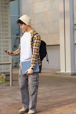 Asian college student checking smartphone on campus plaza holding notebook and backpack
