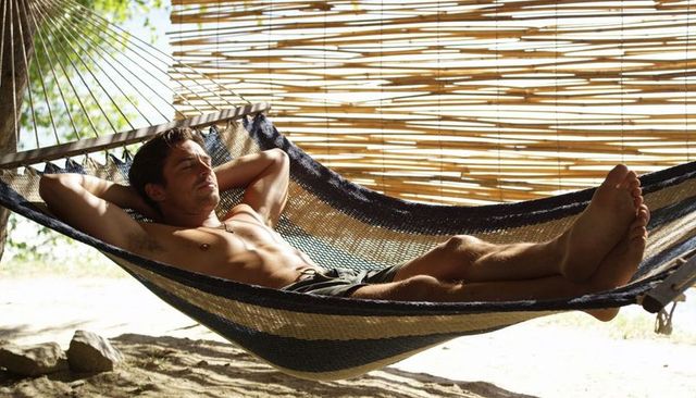Shirtless man relaxing in woven hammock on tropical beach with bamboo privacy screen