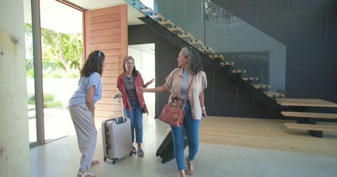 Three women arriving with luggage at modern home foyer greeting host by pivot door