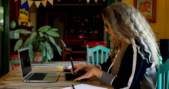 Woman with curly hair using digital tablet and laptop in home office. Perfect for illustrating concepts of remote work, freelance careers, productivity at home, modern work environments, and home office setups.