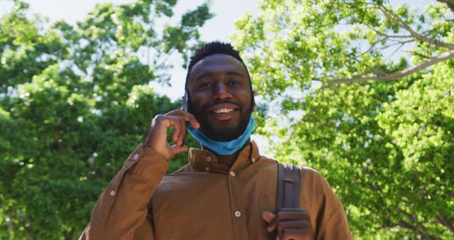 A man in a casual shirt is talking on his smartphone while walking outside with trees and greenery in the background. He is smiling and has a facemask hanging loosely around his neck, indicating a relaxed post-pandemic environment. This image could be used for topics like communication, leisure, technology, post-COVID lifestyle, and outdoor activities.
