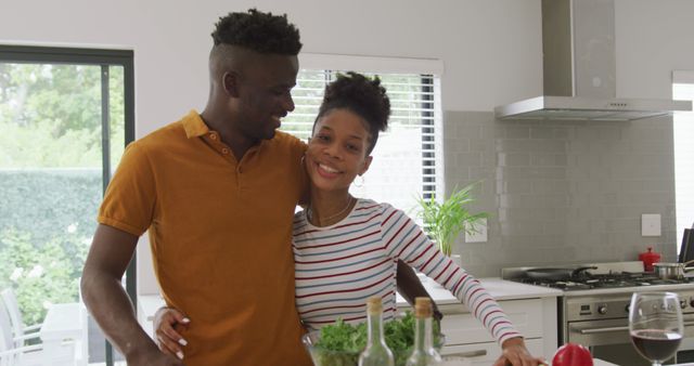 Young African American couple enjoying a moment together while preparing a meal in their modern kitchen. The couple is smiling affectionately and appear to be happy and relaxed. Use this image for content related to relationships, lifestyle, cooking, home life, or healthy living.